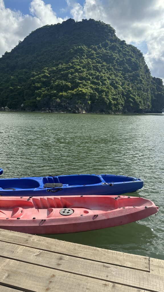 Image of several kayaks in Vietnam. The kayaks are floating in the turquoise waters, surrounded by limestone karsts. The sky is blue and the sun is shining.