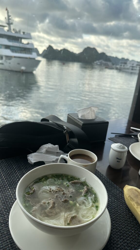 Image of a bowl of Vietnamese soup on a table of a cruise in Ha Long Bay, Vietnam. The soup is a clear broth with noodles, vegetables, and meat. It is served with a side of herbs and spices. The table is set with other dishes, including rice, fruit, and drinks.